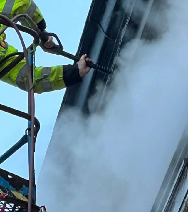 Worker using a high‑pressure washer to clean the exterior wall of a building with visible steam and water spray